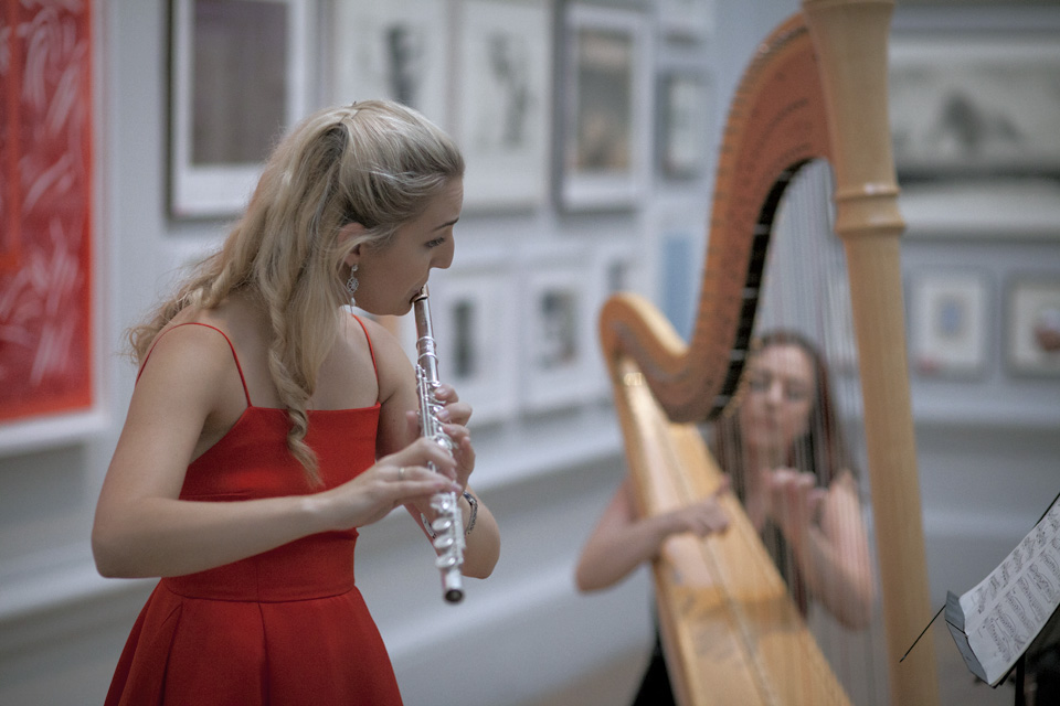 A female flautist, in a red dress, performing the flute, with a female harp performing in the background.